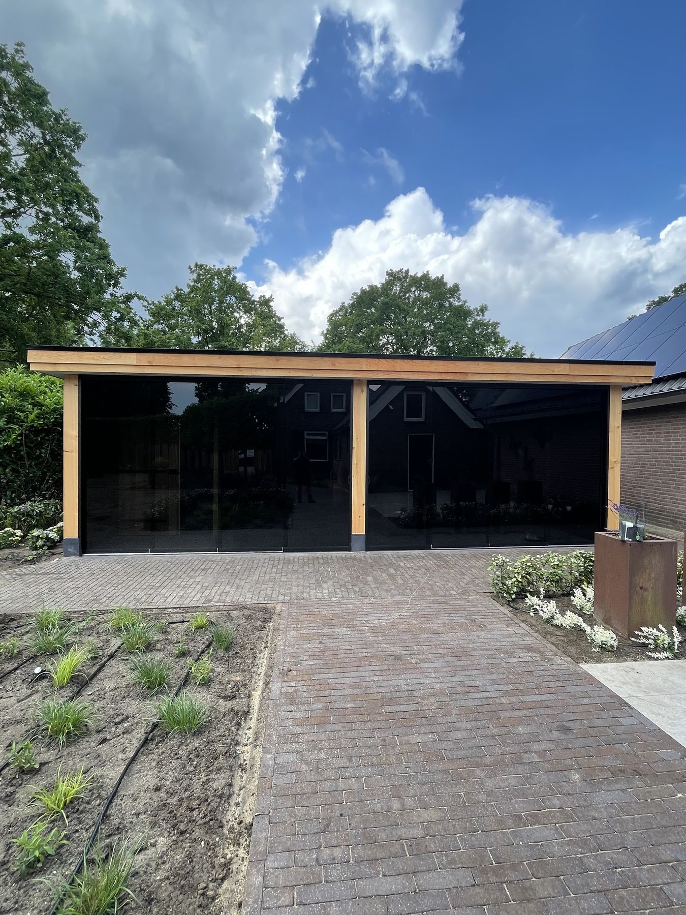 Een lange zwarte houten overkapping met eiken staanders en glazen schuifwanden, met nieuw straatwerk ervoor
