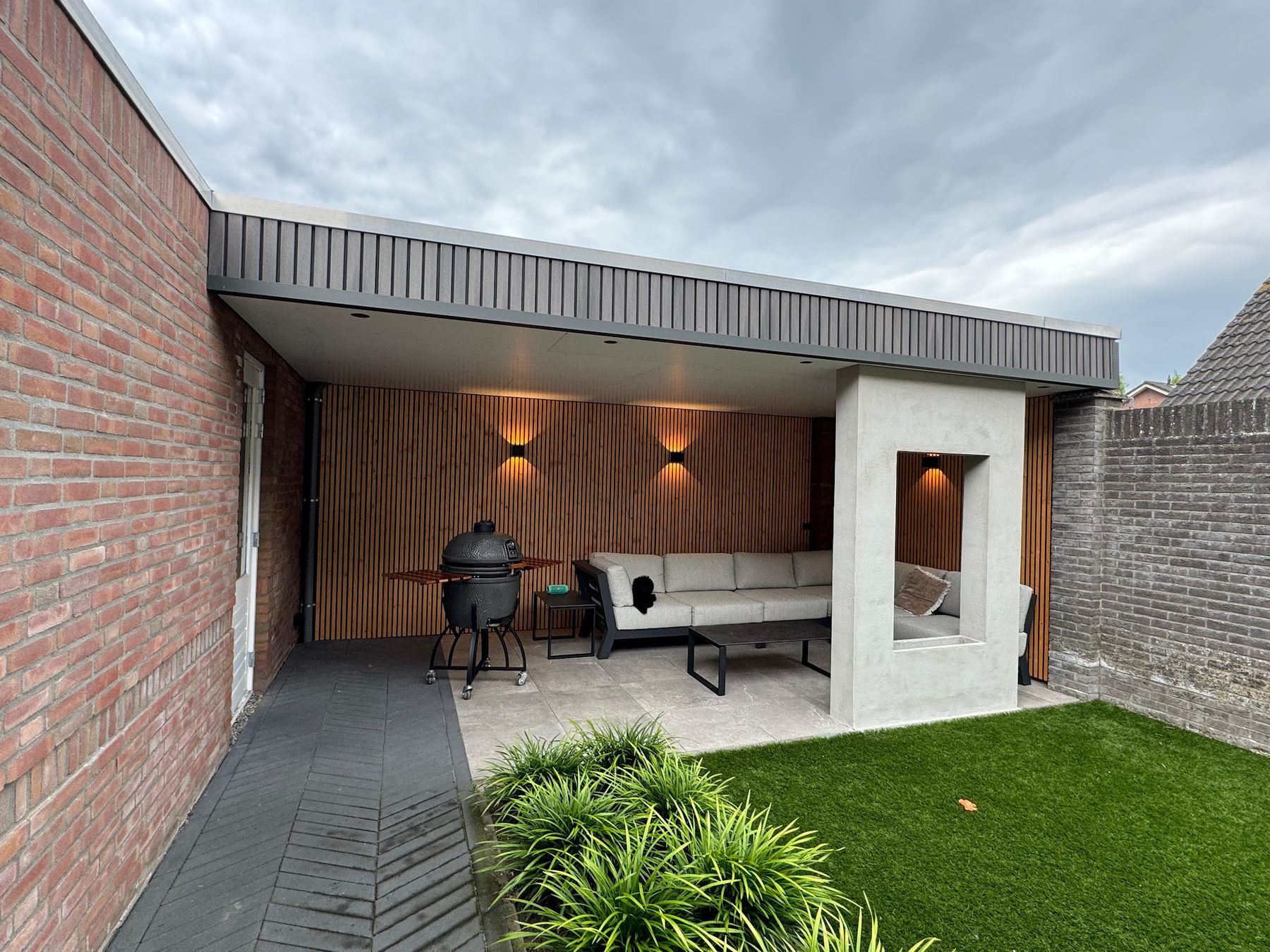 Een moderne overkapping met houten lattenwand, wandlampen, loungebank en barbecue in een tuin met kunstgras