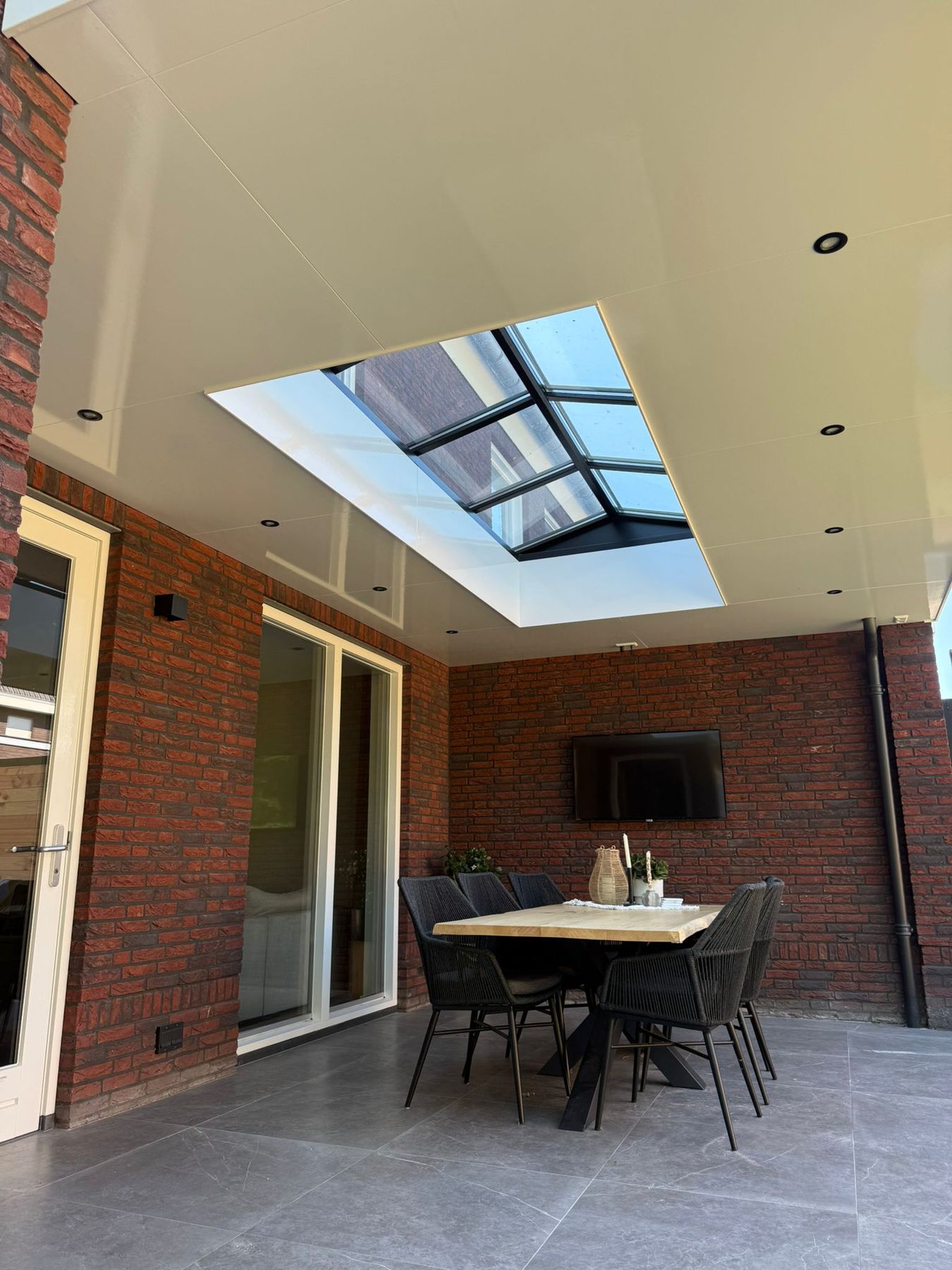 Een afgewerkte veranda tegen een bakstenen gevel met spotjes en een lichtstraat in het witte plafond